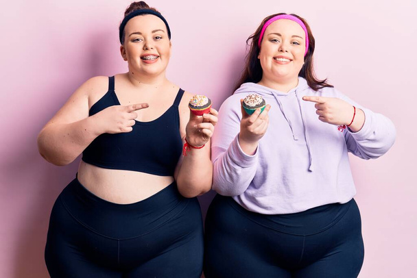 Joven más gemelos de tamaño usando ropa deportiva sosteniendo cupcake sonriendo feliz señalando con la mano y el dedo  - Foto, Imagen