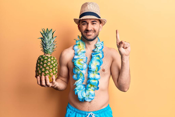 Joven hombre calvo guapo usando traje de baño y lei hawaiano sosteniendo piña sonriendo con una idea o pregunta señalando el dedo con la cara feliz, número uno  - Foto, imagen