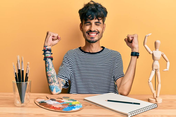 Junger hispanischer Mann, der auf dem Tisch im Kunststudio sitzt, sehr glücklich und aufgeregt und macht Siegergeste mit erhobenen Armen, lächelt und schreit nach Erfolg. Festkonzept.  - Foto, Bild
