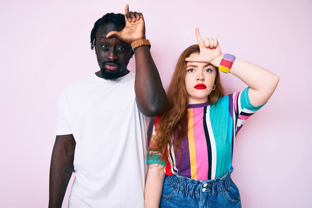 Interracial couple wearing casual clothes making fun of people with fingers on forehead doing loser gesture mocking and insulting.  - Photo, Image