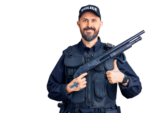 Jeune bel homme portant uniforme de police tenant fusil de chasse souriant heureux et positif, pouce levé faire excellent et signe d'approbation  - Photo, image