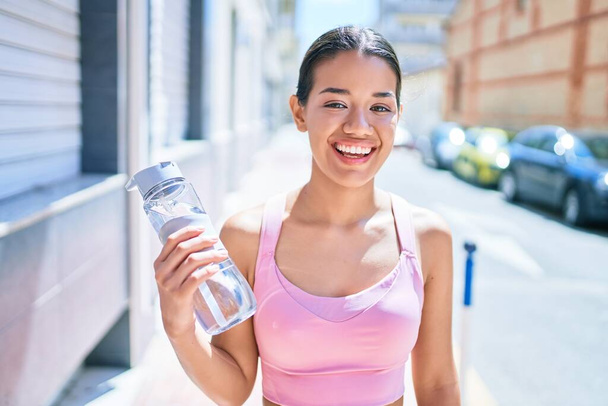 Молодая красивая латиноамериканка в спортивном костюме улыбается счастливой и естественной в городе, пьет свежую воду из бутылки. - Фото, изображение