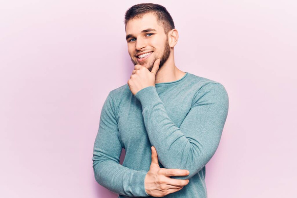 Jonge knappe man draagt casual trui glimlachend zelfverzekerd kijkend naar de camera met gekruiste armen en hand op kin. positief denken.  - Foto, afbeelding