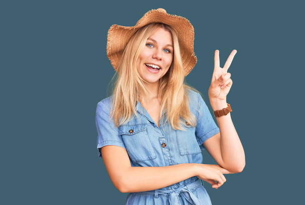 Young beautiful blonde woman wearing summer hat and dress smiling with happy face winking at the camera doing victory sign. number two.  - Photo, Image