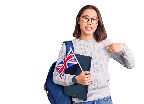 Jeune belle fille chinoise portant un sac à dos étudiant tenant liant et drapeau britannique pointant du doigt vers un sourire heureux et fier  - Photo, image