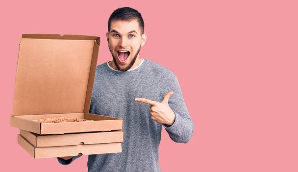 Fiatal jóképű férfi kezében szállítás olasz pizza dobozok mosolyogva boldog mutogatás kézzel és ujjal  - Fotó, kép
