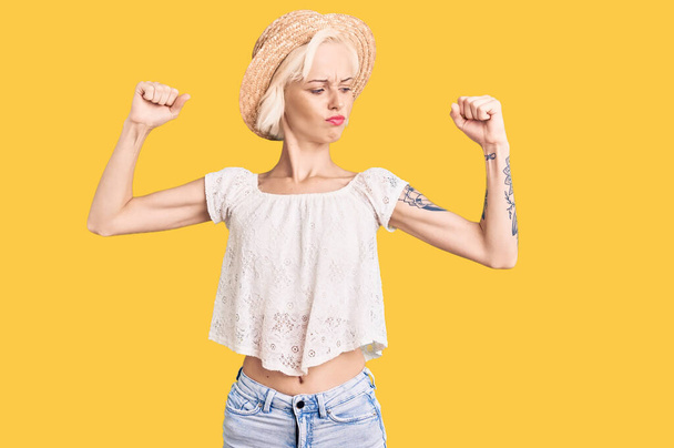 Young blonde woman with tattoo wearing summer hat showing arms muscles smiling proud. fitness concept.  - Valokuva, kuva