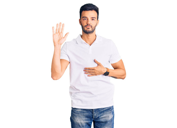 Joven hombre hispano guapo usando ropa casual jurando con la mano en el pecho y la palma abierta, haciendo un juramento de lealtad promesa  - Foto, Imagen