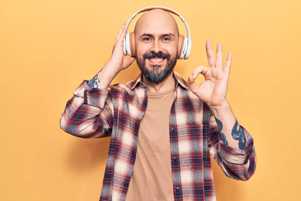 Młody przystojny mężczyzna słuchający muzyki za pomocą słuchawek robi ok znak palcami, uśmiechając przyjazny gest doskonały symbol  - Zdjęcie, obraz