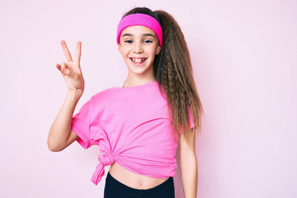 Schattig Latijns-Amerikaans kind meisje draagt sportkleding glimlachend met een vrolijk gezicht knipogen naar de camera doet overwinning teken. nummer twee.  - Foto, afbeelding