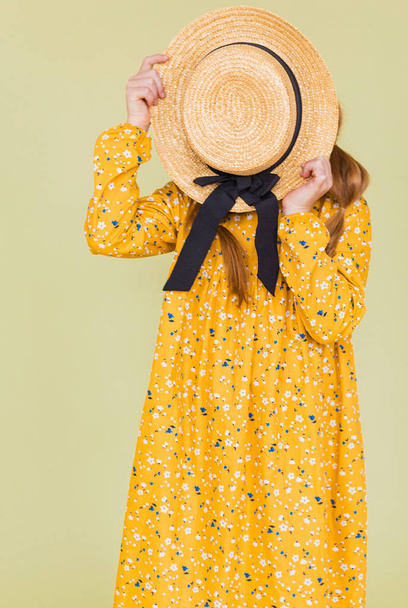 Chica en vestido de lino amarillo posando sobre fondo verde, cubriendo su cara con sombrero de paja. Foto vertical - Foto, imagen