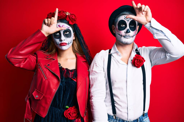 Couple wearing day of the dead costume over red making fun of people with fingers on forehead doing loser gesture mocking and insulting.  - Photo, Image