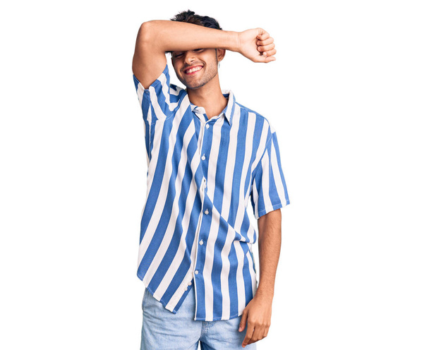Νεαρός Ισπανός που φοράει καθημερινά ρούχα που καλύπτουν τα μάτια με το χέρι χαμογελώντας χαρούμενα και αστεία. τυφλή έννοια.  - Φωτογραφία, εικόνα
