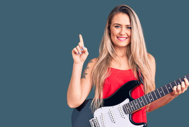 Elektro gitar çalan genç ve güzel sarışın bir kadın bir fikre ya da soru işaretine şaşırdı ve mutlu bir yüzle işaret etti.  - Fotoğraf, Görsel