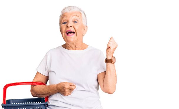 Senior beautiful woman with blue eyes and grey hair holding supermarket shopping basket screaming proud, celebrating victory and success very excited with raised arms  - Photo, Image