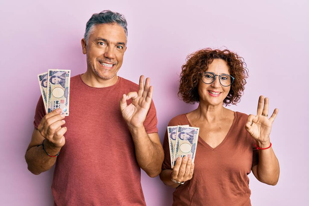Beautiful middle age couple holding 5000 japanese yen banknotes doing ok sign with fingers, smiling friendly gesturing excellent symbol  - Fotoğraf, Görsel