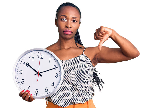 Young african american woman holding big clock with angry face, negative sign showing dislike with thumbs down, rejection concept  - Foto, Bild