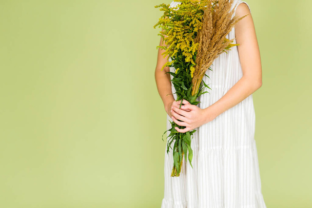 Tyttö valkoisessa pellavamekossa poseeraa vihreällä taustalla kukkakimpun kanssa. Vaakasuora kuva - Valokuva, kuva