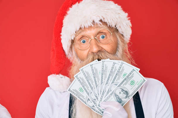 Старик с седыми волосами и длинной бородой в костюме Санта-Клауса держит в руках доллары, празднуя достижение с счастливой улыбкой и выражением победителя с поднятой рукой  - Фото, изображение