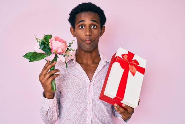 African handsome man holding anniversary present and bouquet of flowers making fish face with mouth and squinting eyes, crazy and comical.  - Photo, image