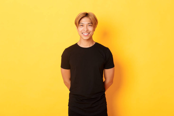 Retrato de amigável bonito asiático cara com cabelo loiro, sorrindo educadamente, de mãos dadas atrás de costas, de pé fundo amarelo - Foto, Imagem
