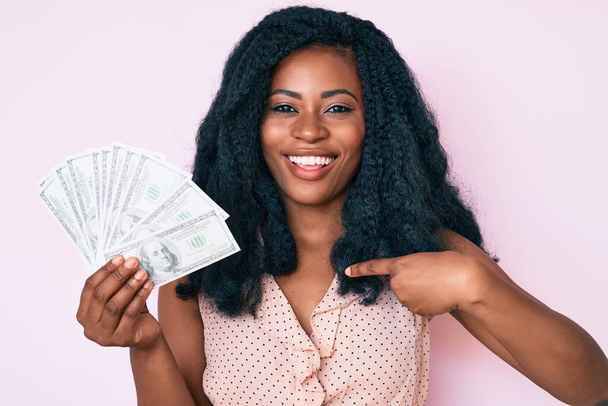 Красивая африканская женщина, держащая доллары, указывая пальцем на себя, улыбается счастливой и гордой  - Фото, изображение