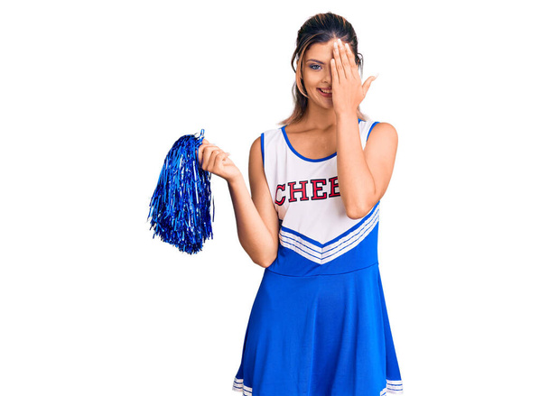 Jonge mooie vrouw dragen cheerleader uniform bedekken een oog met de hand, zelfverzekerde glimlach op het gezicht en verrassing emotie.  - Foto, afbeelding