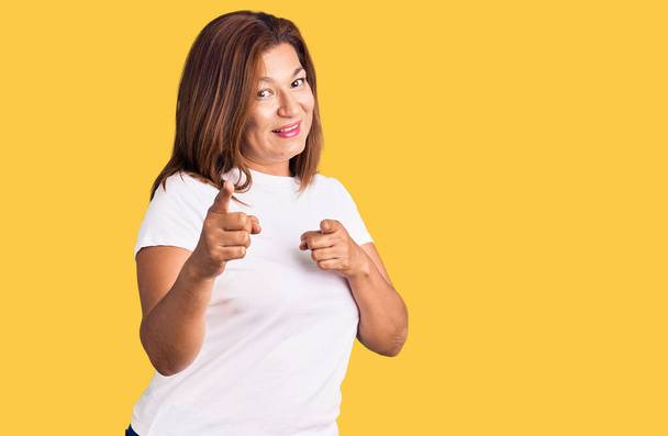 Латиноамериканка средних лет в обычной белой футболке указывает пальцем на камеру со счастливым и смешным лицом. хорошая энергия и вибрации.  - Фото, изображение