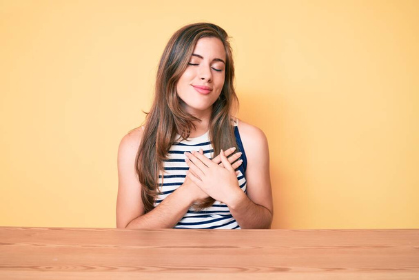 Schöne junge kaukasische Frau in lässiger Kleidung sitzt auf dem Tisch und lächelt mit den Händen auf der Brust mit geschlossenen Augen und dankbarer Geste im Gesicht. Gesundheitskonzept.  - Foto, Bild