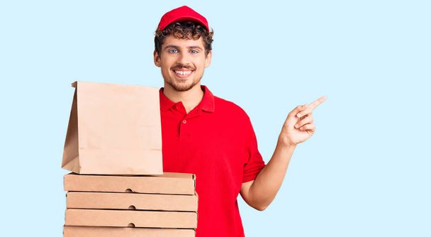 Młody przystojny mężczyzna z kręconymi włosami trzymający pudełko z pizzą uśmiechając się szczęśliwy wskazując ręką i palcem na bok  - Zdjęcie, obraz