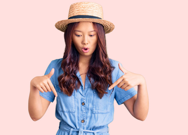 Young beautiful chinese girl wearing summer hat pointing down with fingers showing advertisement, surprised face and open mouth  - Fotografie, Obrázek