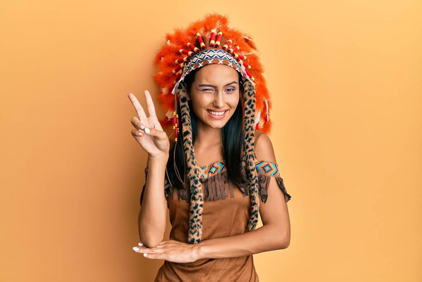 Mladá brunetka v indiánském kostýmu, usmívající se se šťastným obličejem, mrkající na kameru a předvádějící znamení vítězství. číslo dvě.  - Fotografie, Obrázek