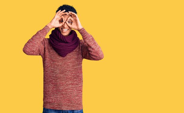 Young african american man wearing casual winter sweater and scarf doing ok gesture like binoculars sticking tongue out, eyes looking through fingers. crazy expression.  - Photo, Image
