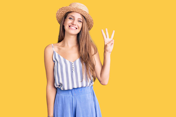 Jovem mulher loira bonita usando chapéu de verão mostrando e apontando para cima com os dedos número três, sorrindo confiante e feliz.  - Foto, Imagem