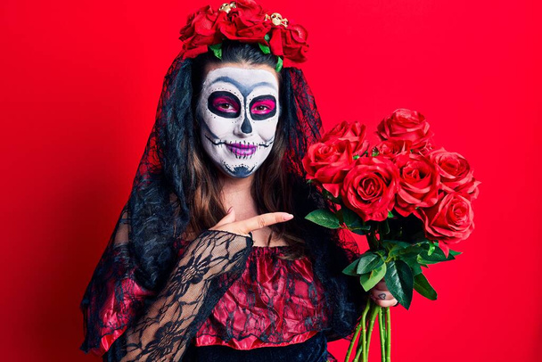 Jonge vrouw draagt dag van het dode kostuum met bloemen glimlachend vrolijk wijzend met hand en vinger  - Foto, afbeelding