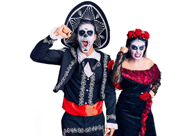 Молодая пара, одетая в мексиканский день мертвого костюма на заднем плане, злая и безумная, поднимающая кулак, разочарованная и разъяренная, крича от гнева. гнев и агрессивная концепция.  - Фото, изображение