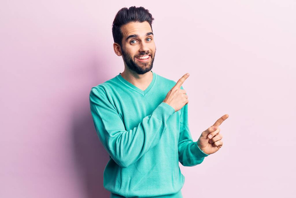 Jonge knappe man met baard in casual sweater lachend en kijkend naar de camera wijzend met twee handen en vingers naar de zijkant.  - Foto, afbeelding
