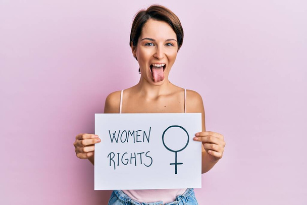 Young brunette woman with short hair holding woman rights banner sticking tongue out happy with funny expression.  - Photo, Image