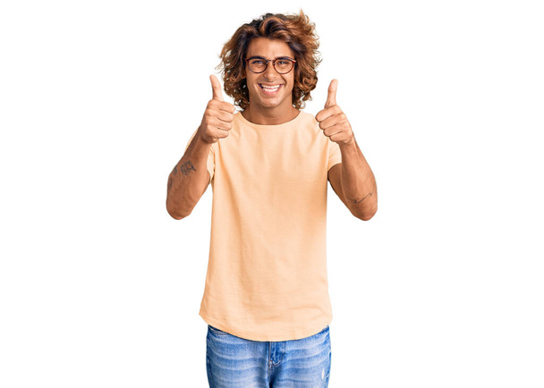 Jonge Spaanse man met casual kleding en bril die positief gebaar met de hand goedkeurt, glimlacht lachend en blij voor succes. winnaar gebaar.  - Foto, afbeelding