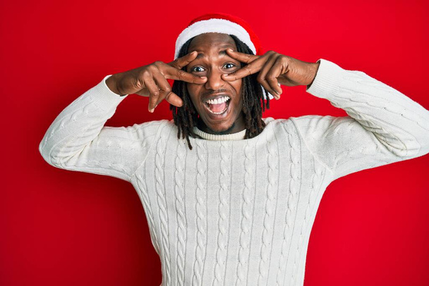 Örgülü Afrikalı Amerikalı adam, Noel şapkası takıyor, barış sembolü takıyor, yüzünde parmaklarıyla gülümsüyor, zafere gülümsüyor.  - Fotoğraf, Görsel