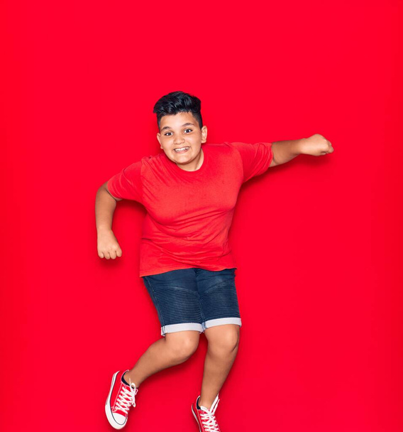 Adorable kid wearing casual clothes smiling happy. Jumping with smile on face over isolated red background - Foto, Bild