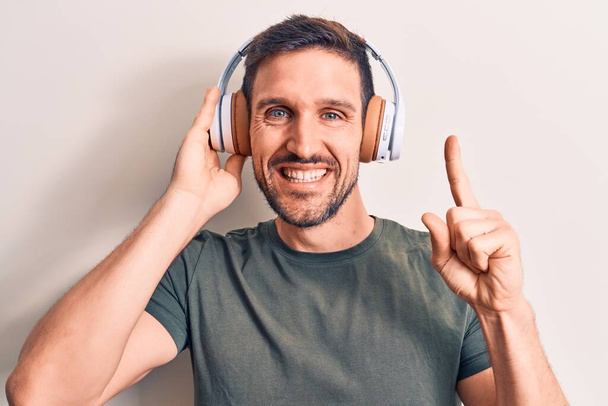 Молодой красивый мужчина слушает музыку, используя наушники на изолированном белом фоне, улыбаясь с идеей или вопросом, указывая пальцем со счастливым лицом, номер один - Фото, изображение