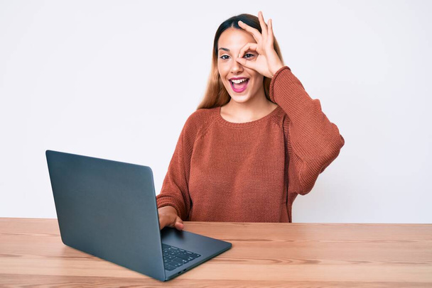 Mujer morena joven trabajando con computadora portátil sonriendo feliz haciendo signo de ok con la mano en el ojo mirando a través de los dedos  - Foto, Imagen
