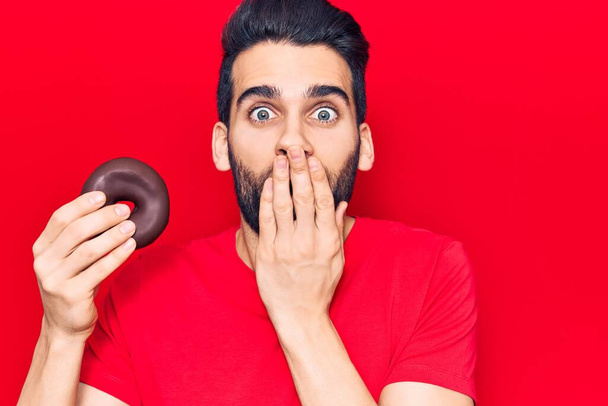 Jonge knappe man met baard die donut met de hand over zijn mond houdt, geschokt en bang voor fouten. verraste uitdrukking  - Foto, afbeelding