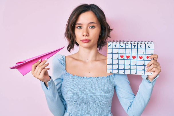 Young beautiful woman holding heart calendar and paper airplane relaxed with serious expression on face. simple and natural looking at the camera.  - Фото, изображение