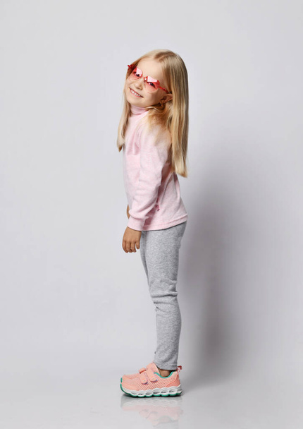 Side view portrait of blonde kid girl in stylish smoked heart-shaped sunglasses, turtleneck shirt sweater and pants - Φωτογραφία, εικόνα