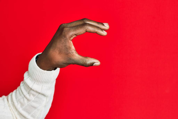 アフリカ系アメリカ人の黒人青年の腕と手で、赤い隔離された背景のピッキングと目に見えないものを取り、指で空間を示すオブジェクトを保持する  - 写真・画像