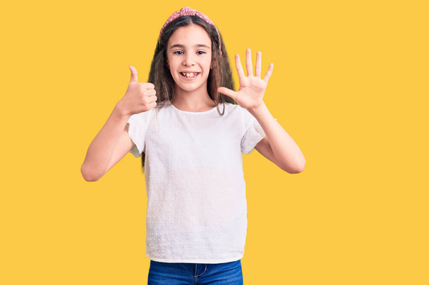 Linda niña hispana usando camiseta blanca casual mostrando y apuntando hacia arriba con los dedos número seis mientras sonríe confiada y feliz.  - Foto, imagen