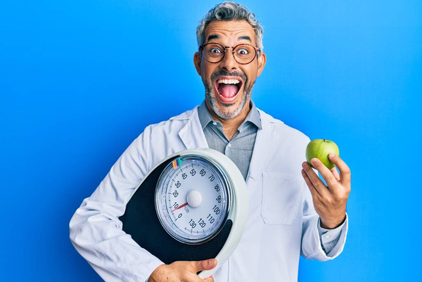 Homme aux cheveux gris d'âge moyen comme médecin nutritionniste tenant la machine de pesage et pomme verte célébrant fou et étonné pour le succès avec les yeux ouverts hurlant excité.  - Photo, image
