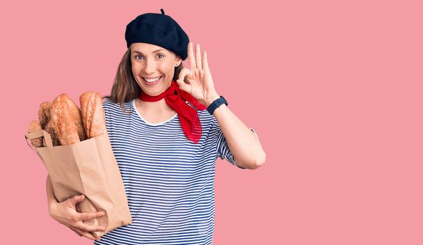 Young beautiful blonde woman wearing french beret holding bag with bread doing ok sign with fingers, smiling friendly gesturing excellent symbol  - Photo, Image